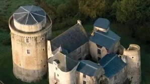 Un bâtiment historique en pierre, doté d’une tour ronde et de plusieurs structures communicantes, le tout entouré de verdure.