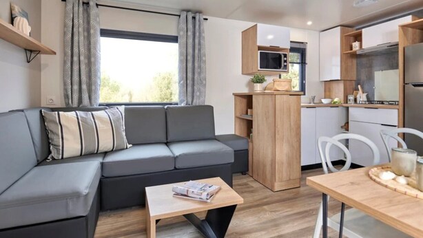 Un espace de vie moderne comprenant un canapé d’angle gris, une table basse en bois, un coin cuisine avec un four à micro-ondes et des placards, ainsi qu’une table à manger avec des chaises.