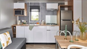 A compact kitchen with white cabinets, a microwave, a gas stove, and a refrigerator. There is a dining area with a table and chairs, and a gray sofa with a patterned pillow. - ZEcamping 3 étoiles pet friendly locatif TV LV clim 6 pers (Pouzauges)