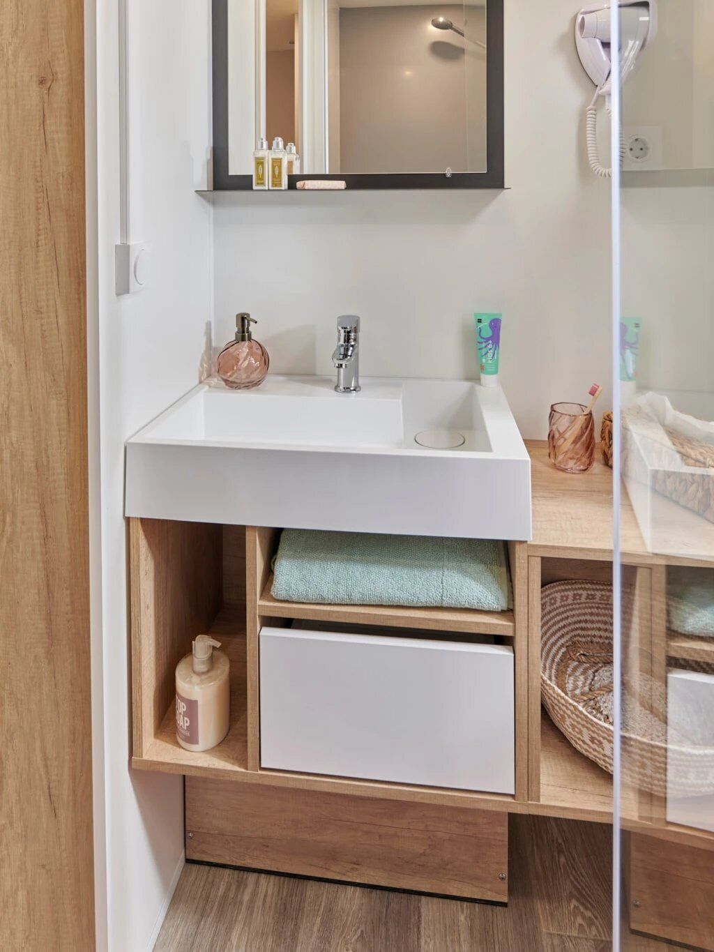Ein modernes Badezimmer mit einem weißen Waschbecken, einem Spiegel und einem an der Wand montierten Fön. Auf dem Waschbecken liegen Pflegeprodukte und auf der Ablage ein Handtuch.