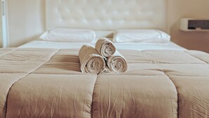 Un lit bien fait, avec une couette et des oreillers de couleur beige, une tête de lit blanche et une table de chevet sur laquelle se trouve un appareil blanc.