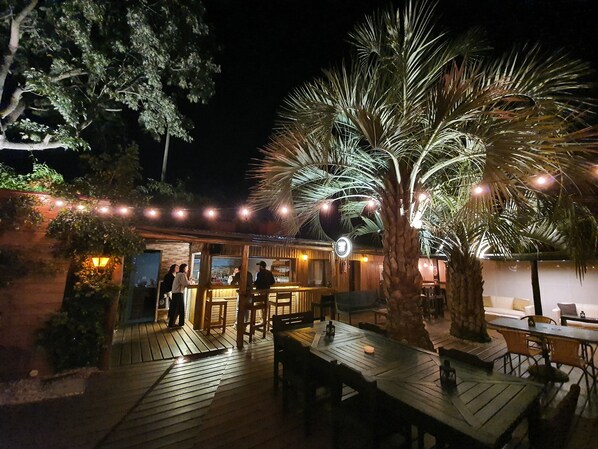An outdoor bar area with wooden tables and chairs, a palm tree, and string lights. - RocaMar Hostel Boutique (Punta del Este)