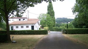 Un bâtiment blanc avec un toit recouvert de tuiles rouges, une allée pavée et une entrée bordée d’une haie.