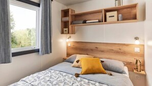 Une chambre à coucher avec un lit, une table de chevet, une étagère en bois au-dessus du lit, une fenêtre avec des rideaux et une vue sur la verdure à l’extérieur.
