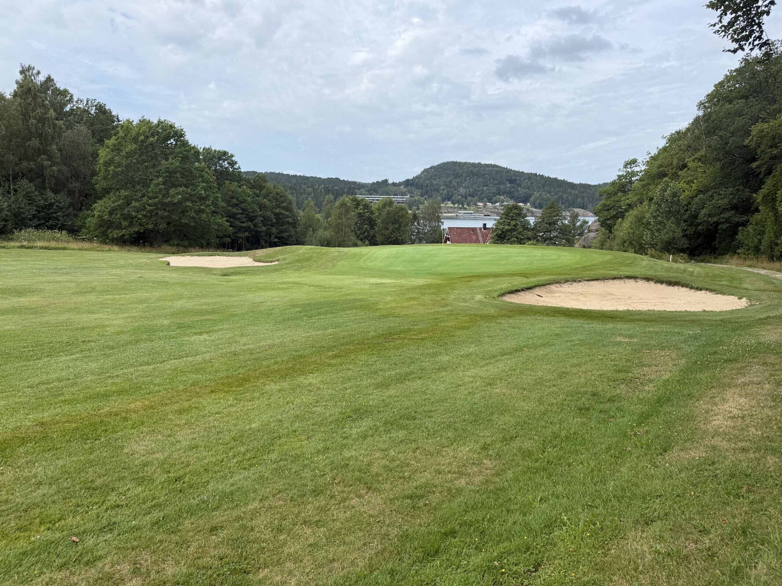 Un parcours de golf avec des bunkers de sable, un green d’entraînement et une vue lointaine sur un bâtiment et des arbres.
