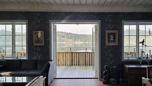 Ein Zimmer mit Blick auf eine bewaldete Landschaft durch große Fenster, eine Holzterrasse und eine Sitzecke mit einer Couch und einem Tisch.