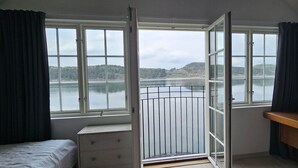 Ein Schlafzimmer mit großem Fenster mit Blick auf einen See, einem Bett, einer Kommode und einem Schreibtisch mit einem gerahmten Bild.