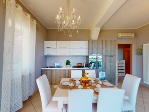 A modern kitchen with white cabinets, a chandelier, and a dining area with white chairs and a table set for a meal. - Agape Apartment by Bookinggardalake (Moniga del Garda)