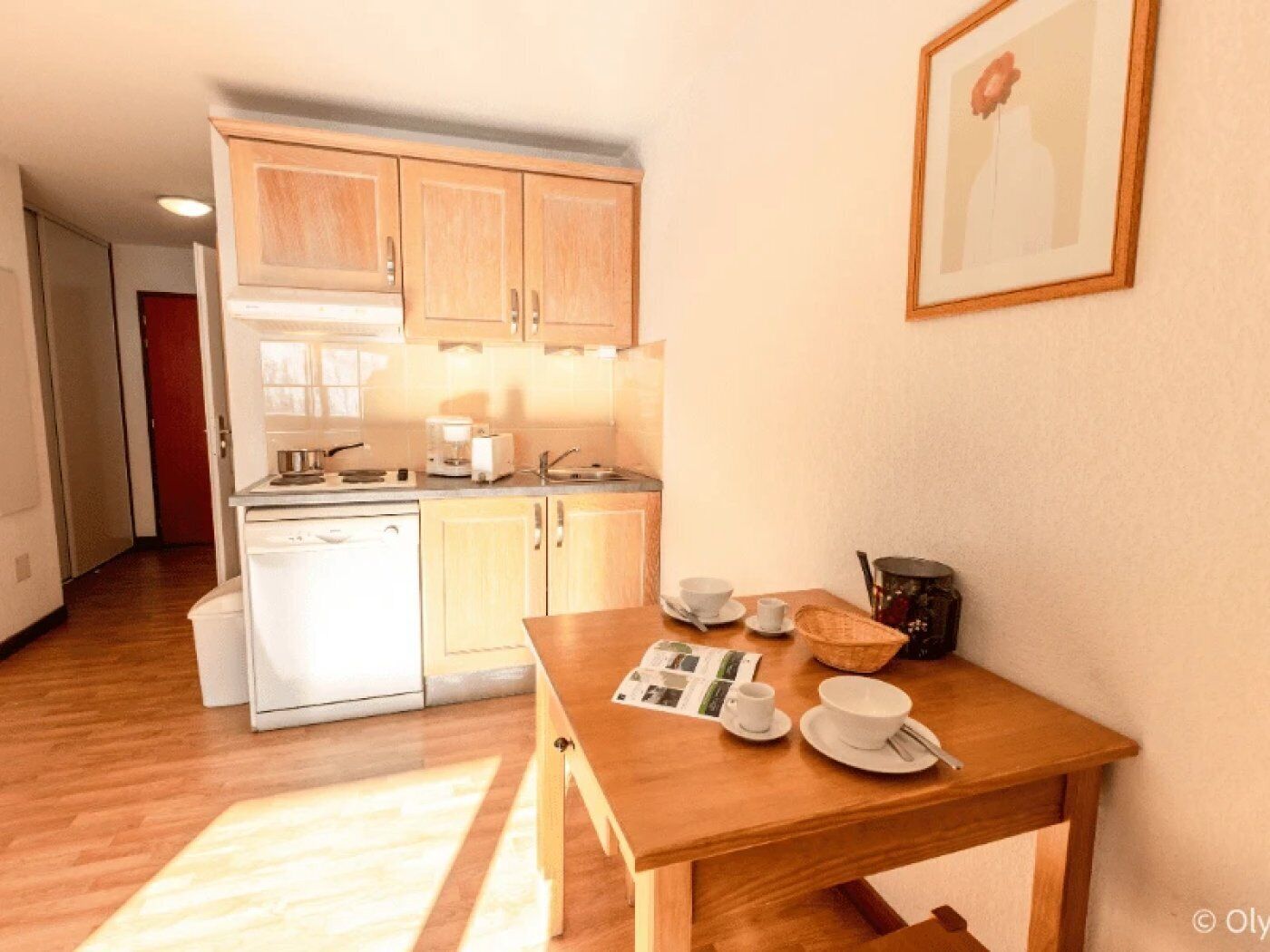 Une cuisine compacte avec des meubles en bois, un lave-vaisselle blanc et une table à manger agrémentée de tasses et d’un saladier.