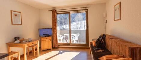 A living room with a sofa, a TV, a wooden table, and a chair. There is a sliding glass door leading to a balcony with outdoor furniture.