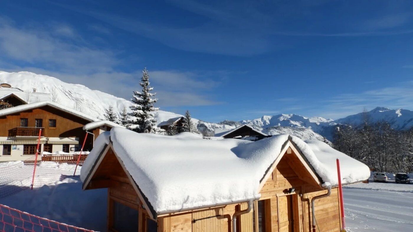 A wooden cabin with a snow-covered roof, surrounded by snow-covered mountains and a clear blue sky.