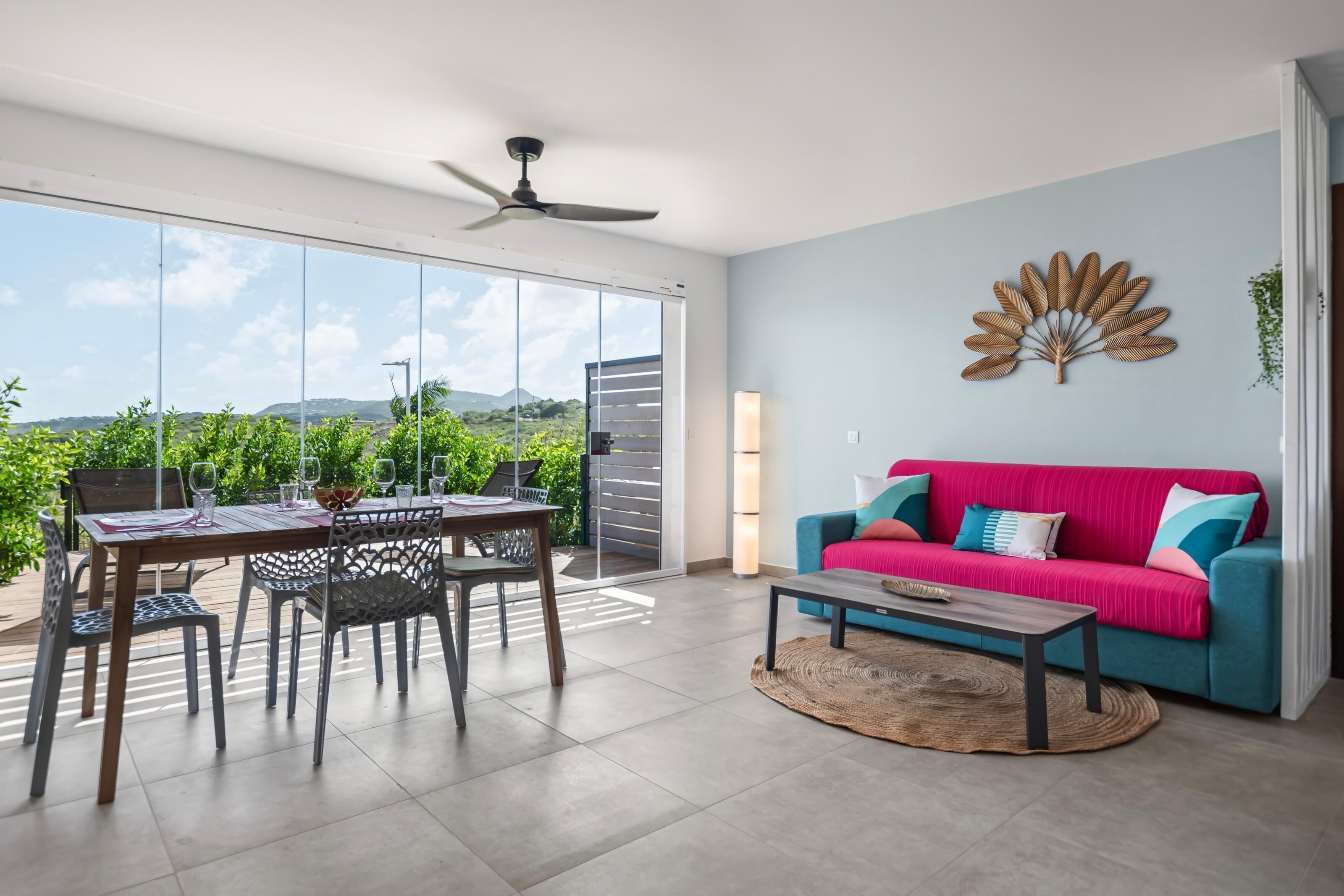 A modern living room with a large sliding glass door leading to a balcony, a bright blue sofa, and a dining area with a table and chairs.