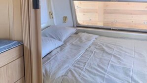 A compact RV interior with a bed, wooden cabinets, and a window providing natural light.