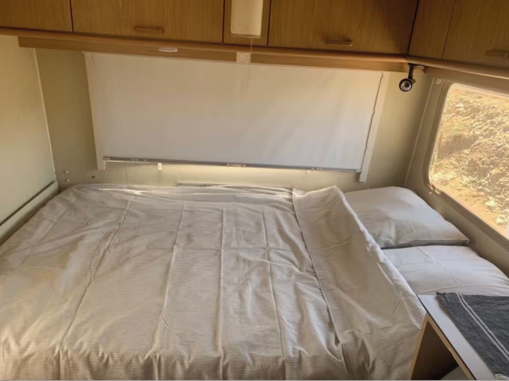 A compact bed with a headboard, a window with a view of the outdoors, and wooden cabinets above the bed.