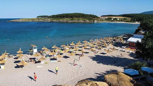 A beach with numerous sun umbrellas and beach chairs, a clear blue sea, and a small hill in the background.