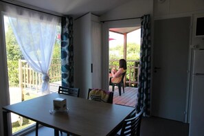 Une pièce avec une table et des chaises, une porte et une vue sur un balcon où une personne est assise à l’extérieur.