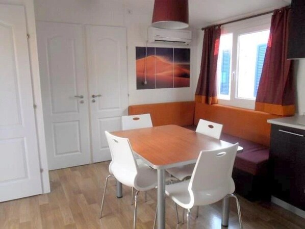 Une pièce de taille modeste comprenant une table à manger en bois, des chaises blanches, une fenêtre avec des rideaux, un appareil de climatisation et un coin cuisine.