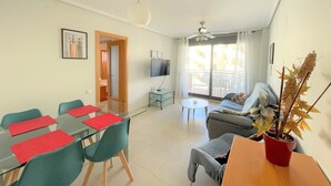 Una sala de estar moderna con una mesa de comedor de vidrio, sillas color esmeralda, un sofá gris y una planta en maceta roja.