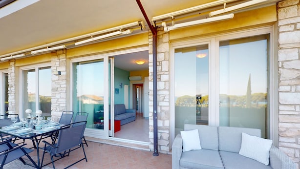 A covered outdoor area with a dining table and chairs, a sofa, and a view of the interior through glass doors. - Horizon luxury lake view by Bookinggardalake (Moniga del Garda)