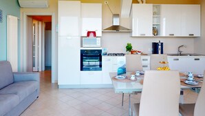 A modern kitchen with white cabinets, a microwave, oven, and a gas stove. There is a dining area with a table set for two, and a sofa in the living area. - Horizon luxury lake view by Bookinggardalake (Moniga del Garda)