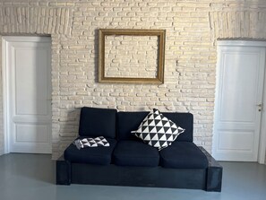 A room with a white brick wall, a black sofa with patterned cushions, and a gold-framed picture frame.