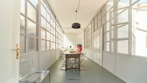 A long, narrow room with a high ceiling, large windows, a central table with chairs, and a hanging light fixture.