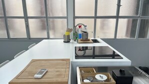 A modern kitchen with a white countertop, a wooden cutting board, a sink with various kitchen items, and a window with a view of buildings.