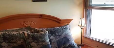 Una cama con cabecera, dos almohadas decorativas, una mesita de noche con lámpara y una ventana con persianas.