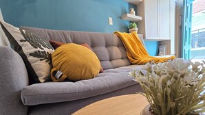 Un sofá gris con dos almohadas color mostaza y una almohada decorativa con forma de hoja. Una manta amarilla está colocada sobre el sofá. Una planta en maceta con flores blancas se encuentra sobre una mesa de madera en primer plano.