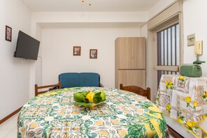 Una habitación con cama, televisor montado en la pared, ventana con persianas y una mesa con mantel estampado.