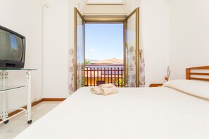 Een hotelkamer met een bed, een televisie op een trolley, en uitzicht op een gebouw met rode dakpannen door een geopende deur.
