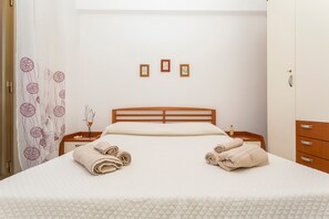 Una cama bien hecha con toallas enrolladas, un cabecero de madera, una mesita de noche con una botella y una pared con tres cuadros enmarcados.