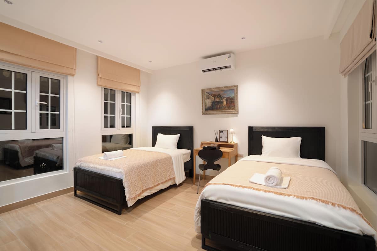 A hotel room with two single beds, a desk, a chair, and a window with blinds.