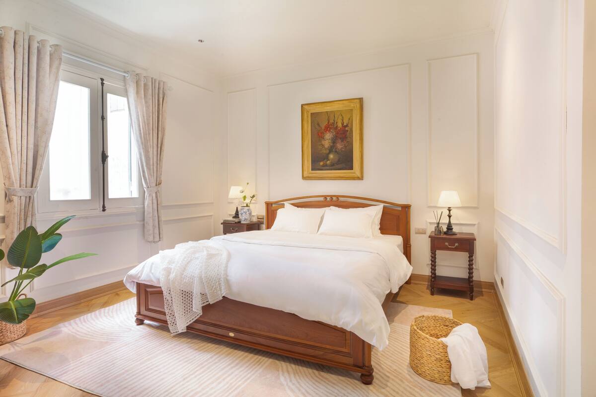 A neatly arranged bedroom with a wooden bed, two bedside tables with lamps, a large window with curtains, and a painting on the wall.