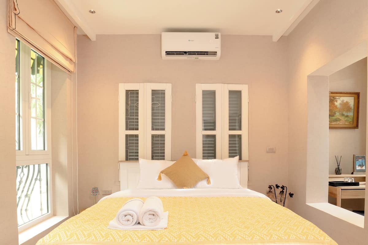 A bedroom with a large bed, white bedding, a window with blinds, and an air conditioning unit on the wall.