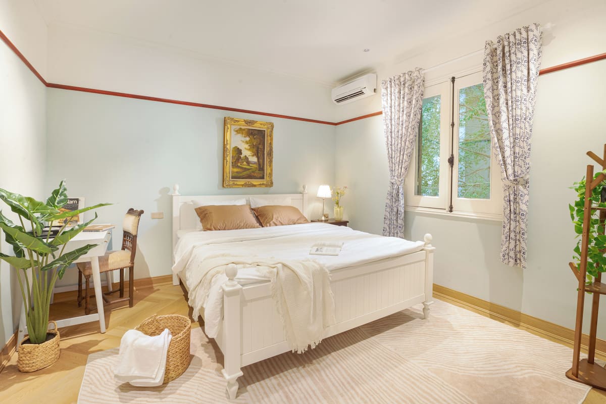 A bedroom with a bed, bedside table, lamp, window with curtains, and a painting on the wall.