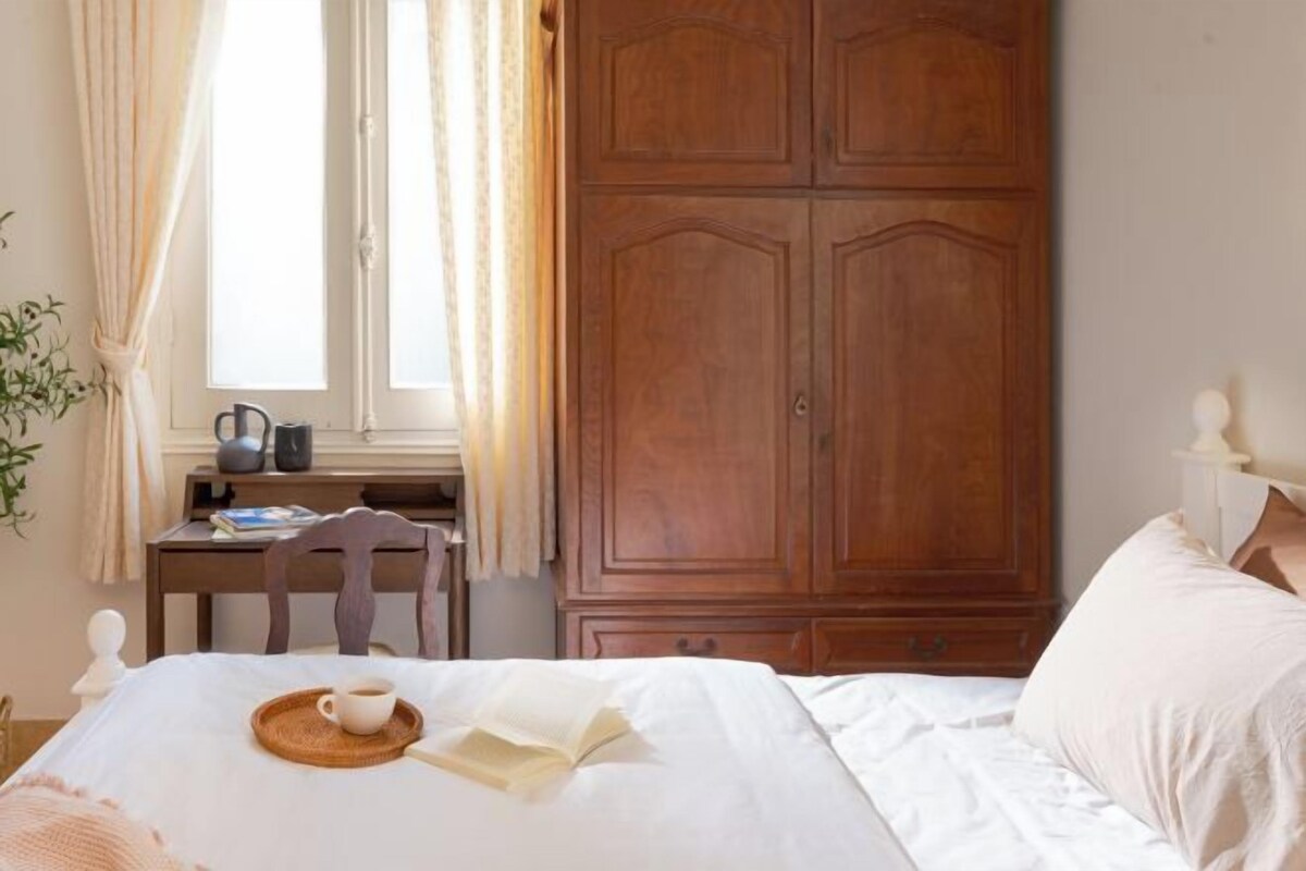 A bedroom with a bed, a wooden wardrobe, a desk with a kettle, and a window with curtains.