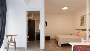 Un dormitorio con cama, una silla, una mesita y un baño visible a través de una puerta abierta.