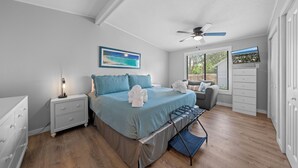 Une chambre à coucher comprenant un lit, des tables de chevet, un ventilateur de plafond, une fenêtre avec des stores et un téléviseur.