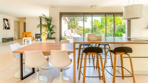 A modern kitchen with a large island, white chairs, and a view of greenery outside.