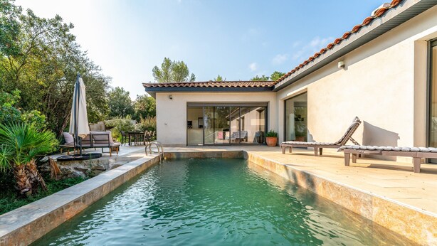 A swimming pool with a patio area, outdoor furniture, and a building with large windows.