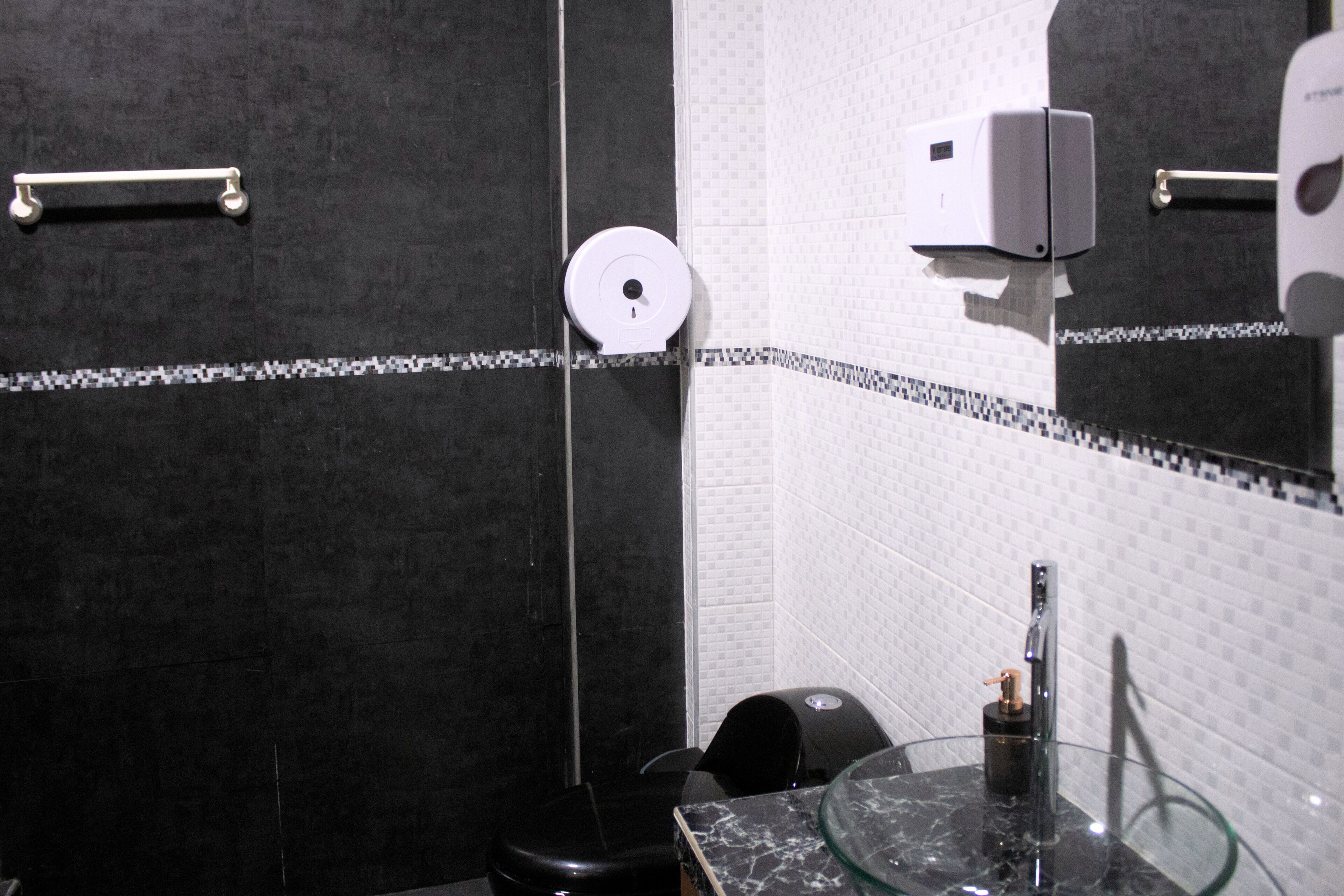 Un baño moderno con lavamanos de vidrio, inodoro negro, dispensador de toallas de papel y dispensador de jabón.