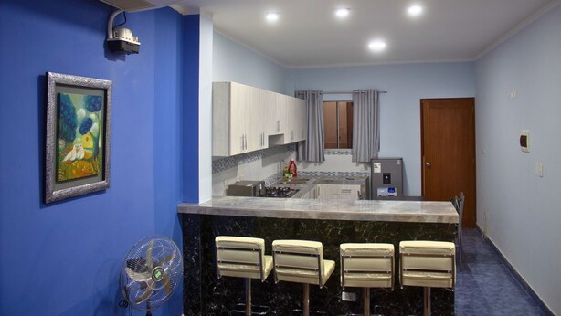 A compact kitchen with a marble countertop, a sink, and a refrigerator. There are three bar stools and a ceiling fan.