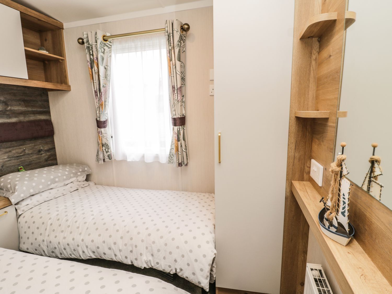 Une chambre avec une tête de lit en bois, une fenêtre ornée de rideaux à motifs, un lit avec une literie à pois et une étagère en bois sur laquelle sont disposés des objets décoratifs.
