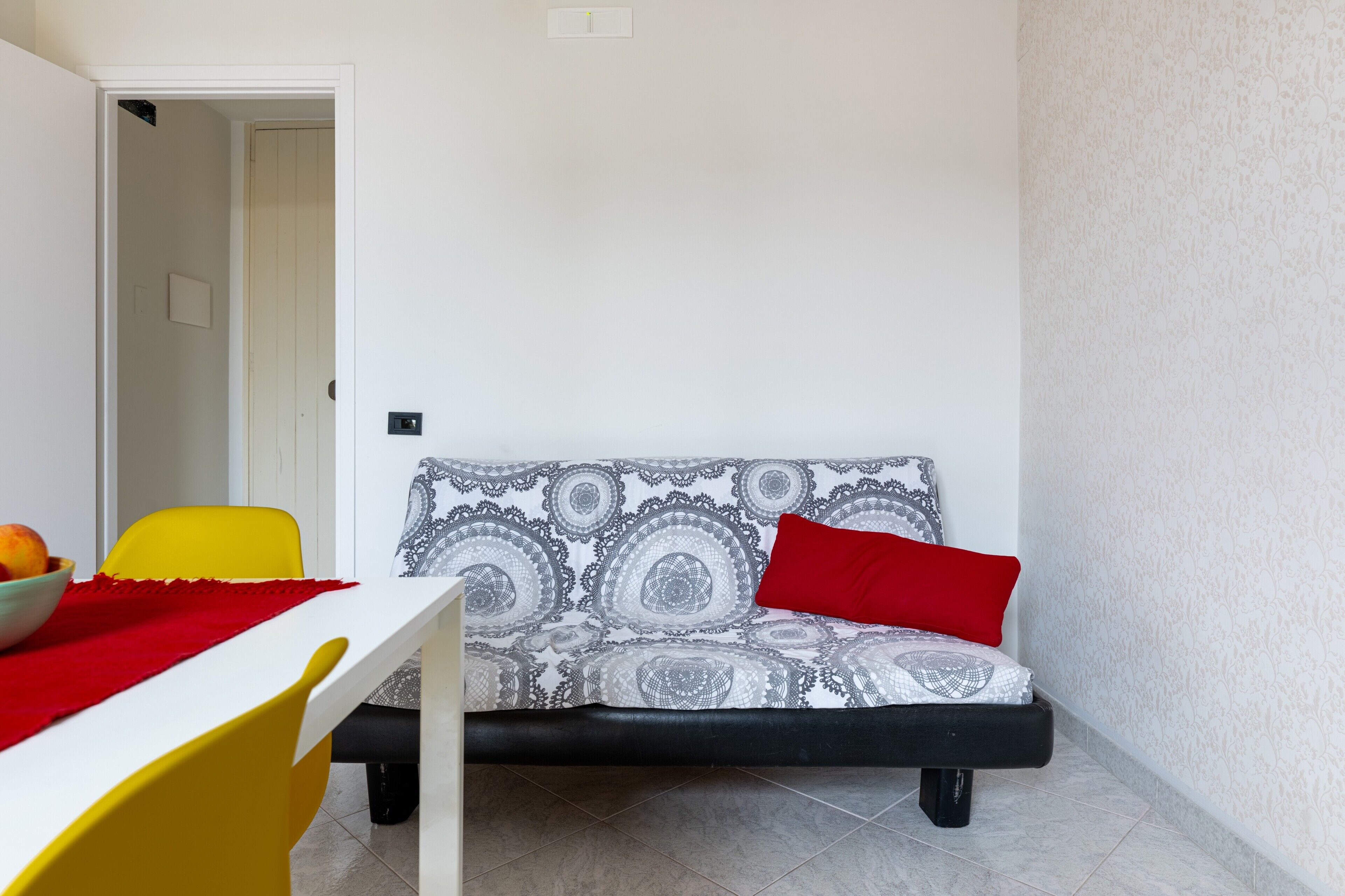 Ein Zimmer mit einem weißen Tisch, einem gelben Stuhl, einem gemusterten Sofa mit rotem Kissen und einer Tür zu einem anderen Raum.