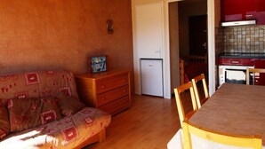 A cozy living room with a wooden ceiling, a sofa with patterned cushions, a small wooden dresser, and a dining table with chairs.