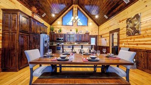 A wooden cabin interior with a dining table set for four, a kitchen with modern appliances, and a large window providing natural light.