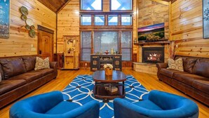 A log cabin interior with a fireplace, two leather sofas, a coffee table, and a large window.