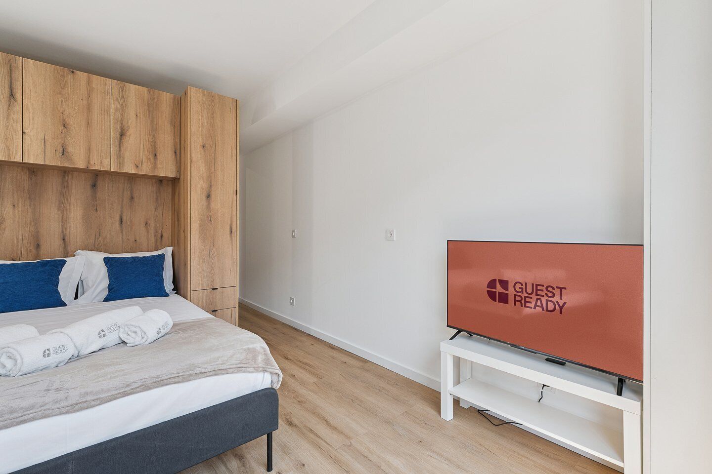 A modern hotel room with a bed, wooden headboard, a flat-screen TV, and a white nightstand.