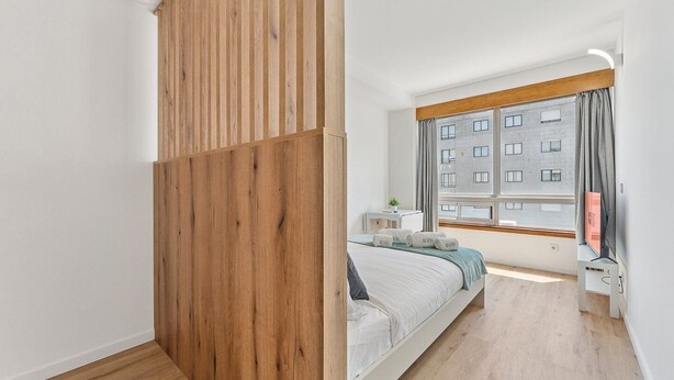 Une chambre moderne avec une cloison en bois, un lit doté d’une tête de lit grise, une table de chevet avec une lampe, et une vue sur un immeuble par la fenêtre.